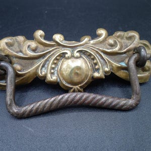 May include: A vintage brass drawer pull with an ornate, scrolled design. The pull has a twisted metal handle.