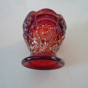 May include: A red glass candy dish with a decorative pattern. The dish has a wide, round base and a flared top.