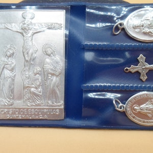 May include: A silver religious artifact set in a blue case. The set includes a rectangular plaque with a depiction of the crucifixion, two oval pendants, and a cross pendant. The plaque has the text "S. CRISTO DE ESQUIPULAS GUATEMALA-C.A."