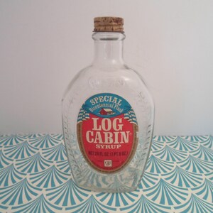 May include: A clear glass bottle with a cork stopper and a red, white, and blue label that reads "SPECIAL Bicentennial Flash LOG CABIN SYRUP NET 24 FL OZ (1 PT. 8 OZ)"