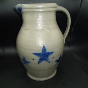 May include: A stoneware pitcher with a handle and a spout. The pitcher is off-white with blue star designs. The rim and spout have blue accents. The pitcher has a bulbous body and a cylindrical neck.
