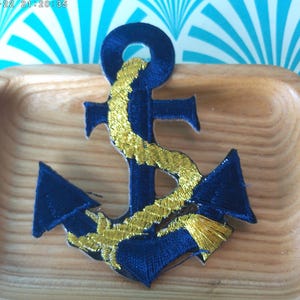May include: A blue and gold embroidered anchor patch. The anchor is a nautical symbol and is often used as a decorative element on clothing and accessories.