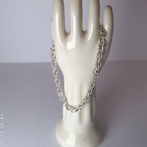 May include: A silver chain bracelet with a double-link design is draped over a white ceramic hand. The bracelet is made of shiny, polished metal. The hand is a decorative display piece, set against a soft, neutral background.