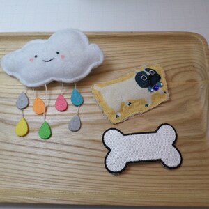 May include: A set of three felt appliques featuring a white cloud with a smiling face and raindrops, a tan pug with a blue jewel, and a white bone.