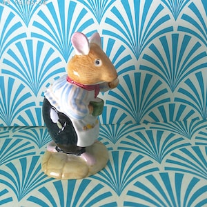 May include: A ceramic figurine of a mouse wearing a blue and white striped shirt, a red scarf, and a white apron. The mouse is holding a green mug. The figurine has a tan body, pink ears and feet, and black pants. It stands on a tan base.