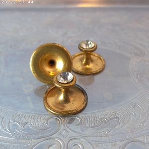 May include: Three vintage gold-toned cufflink studs. Two studs have a small, round, clear rhinestone on top. The third stud has a flared, trumpet-like shape. The cufflinks are displayed on a silver, ornate tray.
