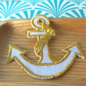 May include: A white anchor with gold embroidery. The anchor is on a wooden surface.
