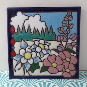 May include: Square ceramic tile with a hand-painted floral and landscape design. The tile features a blue sky, white clouds, green trees, and various flowers in shades of blue, pink, and purple. The tile is framed with a dark blue border.