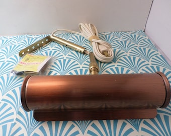 May include: A copper-colored wall-mounted light fixture with a cylindrical shade and a white cord. The light fixture is attached to a white wall with a blue and white patterned design. The light fixture comes with a bracket and instructions.
