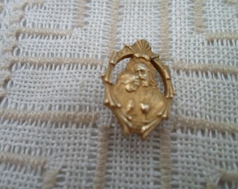 May include: A gold-colored pendant with an oval frame and a religious figure. The pendant features a detailed depiction of a bearded man with a halo, and a heart. The background is a cream-colored, textured fabric.
