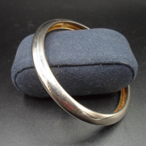 May include: A silver bangle bracelet with a smooth, rounded surface. The bracelet is lying on a dark blue velvet cushion.