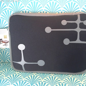 May include: A dark gray laptop sleeve with a gray trim and a geometric design of gray circles and lines. The sleeve has a zipper closure and a tag with an image of a chair and ottoman.