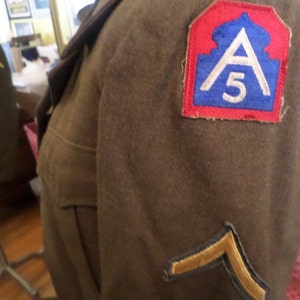 May include: A brown military jacket with a patch on the sleeve that says "A 5" in white lettering on a blue background. The patch is surrounded by a red border. There is a gold chevron on the lower sleeve.