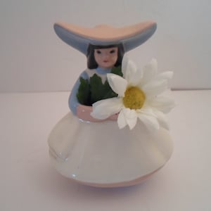 May include: A ceramic figurine of a girl wearing a pink and blue hat and a white dress, holding a white daisy.