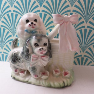 May include: A vintage ceramic planter shaped like two dogs, one white with a blue bow and one black and white with a pink bow. The dogs are sitting in a basket with a pink bow.