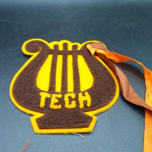 May include: A brown felt patch shaped like a lyre with the word "TECH" in brown felt letters. The patch has a yellow border and is attached to a brown and orange ribbon.