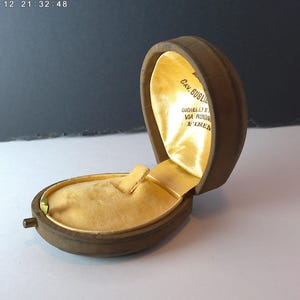 May include: An open, oval-shaped jewelry box with a brown exterior and a gold satin interior. The box is hinged and has a small clasp. The interior has a small, rectangular holder. The text "CAV. GUGLI" and other text is visible inside the lid.