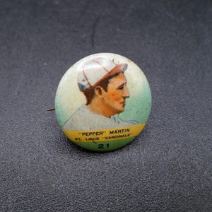 Może przedstawiać: Vintage przypinka baseballowa z portretem "Pepper" Martin z St. Louis Cardinals. Przypinka ma jasnozielone i ż&oacute;łte tło z nazwiskiem gracza i szczeg&oacute;łami drużyny. Widoczny jest r&oacute;wnież numer 21.