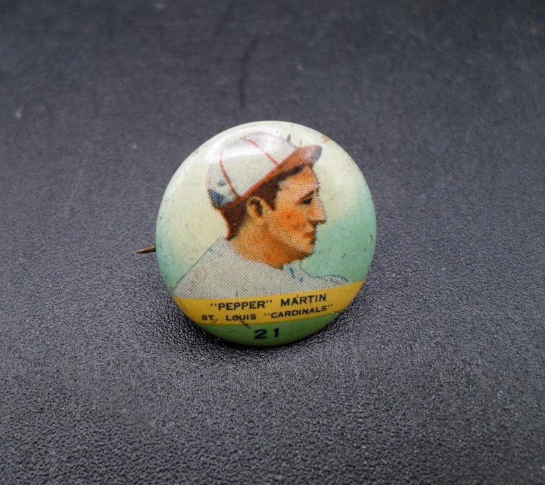 Może przedstawiać: Vintage przypinka baseballowa z portretem "Pepper" Martin z St. Louis Cardinals. Przypinka ma jasnozielone i ż&oacute;łte tło z nazwiskiem gracza i szczeg&oacute;łami drużyny. Widoczny jest r&oacute;wnież numer 21.