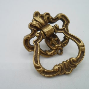 May include: A gold-toned ornate metal drawer pull with a decorative design. The pull has a ring shape with a detailed floral pattern.