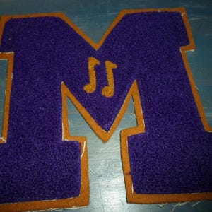 May include: Purple felt letter M with a gold felt outline. The letter M has two gold musical notes inside.