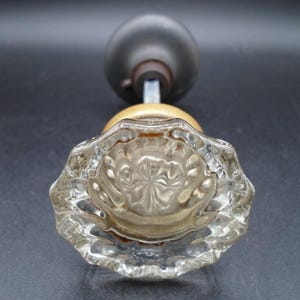 May include: A clear, faceted glass doorknob with a brass base and a dark gray knob. The glass knob has a starburst pattern in the center. The doorknob is likely vintage or antique.