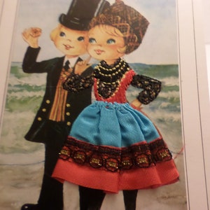 May include: A vintage postcard featuring a couple in traditional folk costumes. The woman wears a blue and red skirt with black lace trim and a beaded necklace. The man wears a black suit with a striped scarf. The postcard has a seaside background.