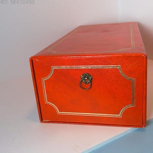 May include: A rectangular orange box with gold trim and a decorative brass ring pull. The box has a textured surface and a gold border around the front panel. The box is likely for storage.