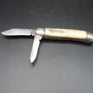 May include: A vintage pocket knife with a bone handle and two blades. The knife has a silver-colored metal frame and a small, engraved design on the handle.