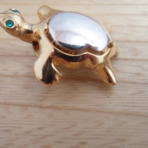 May include: A gold-colored turtle brooch with green gem eyes. The turtle's shell is silver-colored.