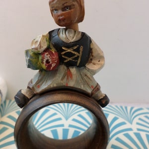 May include: Wooden napkin ring with a hand-painted figurine of a person wearing a traditional costume and holding flowers. The figurine is painted in shades of brown, red, green, and blue. The napkin ring is made of dark wood.