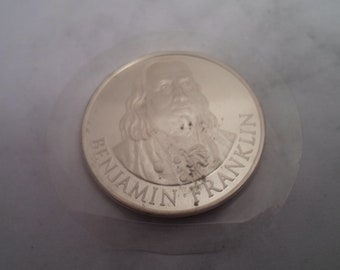 May include: Silver coin featuring a portrait of Benjamin Franklin with the inscription "Benjamin Franklin" around the edge.