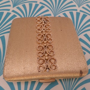 May include: A gold compact with a decorative design on the front. The design is made of a gold-colored metal and features a repeating pattern of circles and loops.
