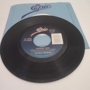 May include: A black vinyl 45 RPM record with a blue and white label. The label has the text "Epic" in a stylized font and the title "Nothin' New" by Gloria Estefan.