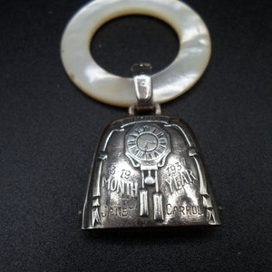 May include: Silver baby rattle with a bell shaped charm. The charm has a clock face with the inscription "1935 YEAR" and "MONTH 1935". The inscription "JANET CARROL" is also on the charm.