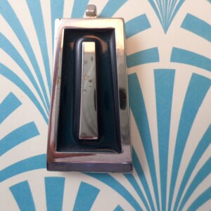 May include: A silver pendant with a blue enamel rectangle and a silver bar inside. The pendant is on a white background with a blue and white pattern.
