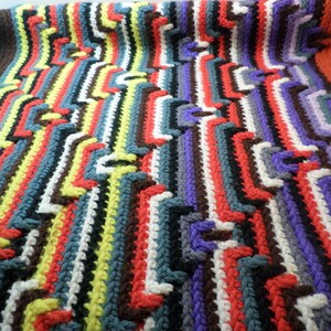 May include: A colorful crocheted blanket with a zig-zag pattern in shades of red, orange, yellow, green, blue, purple, brown, black, and white.
