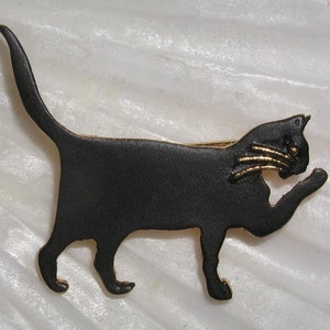 Könnte beinhalten: Schwarze Metall-Katzenbrosche mit goldenen Akzenten an den Ohren und Augen. Die Katze ist in einer Gehposition mit eingerolltem Schwanz.