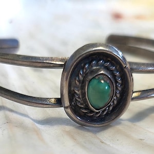 May include: A silver cuff bracelet with an oval turquoise stone set in a decorative bezel. The bracelet has a double-band design and a vintage aesthetic. The turquoise stone is a vibrant green color.