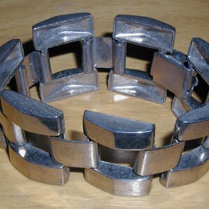 May include: A silver-toned metal bracelet with a geometric design. The bracelet is composed of rectangular links connected by smaller, square-shaped links. The links have a slightly curved shape, and the bracelet is resting on a wooden surface.
