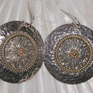 May include: A pair of silver-tone earrings with a hammered texture. Each earring features a central gold-tone floral design surrounded by a gold-tone beaded border. The earrings have a hook closure.