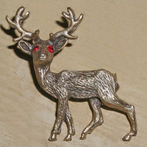 May include: A gold-toned metal deer brooch with red rhinestone eyes. The deer is standing on all four legs with its head held high. The antlers are curved and point upwards.