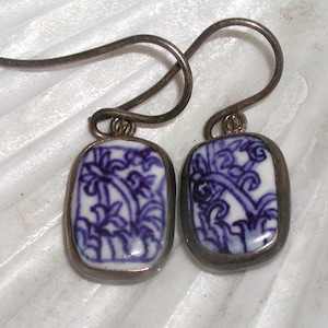 May include: A pair of earrings with rectangular, white ceramic pendants featuring a blue floral design. The pendants are framed in a silver-toned metal and attached to silver-toned ear wires. The earrings have a vintage aesthetic.