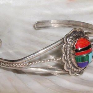May include: A silver bracelet with a multi-colored stone inlay. The stone is set in a silver bezel and has a rope-like design around the edge. The bracelet is made of two bands of silver that are twisted together.
