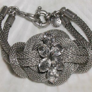 May include: A silver chain bracelet with a knotted design and a clear rhinestone flower accent. The bracelet has a lobster clasp closure.