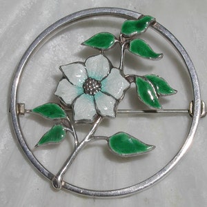 Könnte beinhalten: Eine silberne Brosche mit einer weißen Blume und grünen Blättern in einem Kreis.