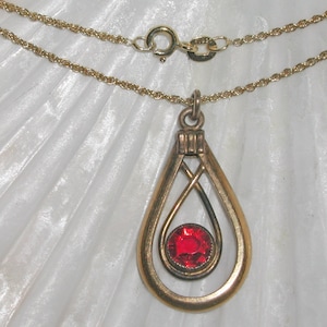 Puede incluir: Un collar de cadena de oro con un colgante en forma de lágrima. El colgante tiene una piedra preciosa roja en el centro y un diseño delicado e intrincado.
