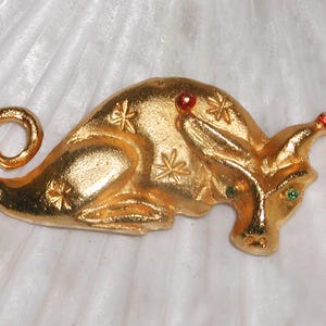 May include: A gold-toned metal pin shaped like a seahorse with red and green gemstones. The seahorse has a curled tail and a star-shaped design on its body.