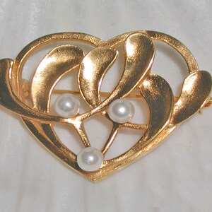 Könnte beinhalten: Eine goldene herzförmige Brosche mit einem filigranen Design aus Blättern und drei weißen Perlen.
