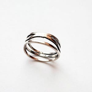 May include: A silver ring with a layered design. The ring features three bands that curve and overlap, creating a textured appearance. The ring is set against a white background, highlighting its metallic shine.